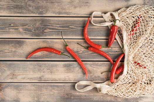 Red hot tasty chili peppers scattered near ecological degradable string bag on wooden background. Spicy food, seasoning and condiment. Sustainable resource, environmental care. Top view, copy space