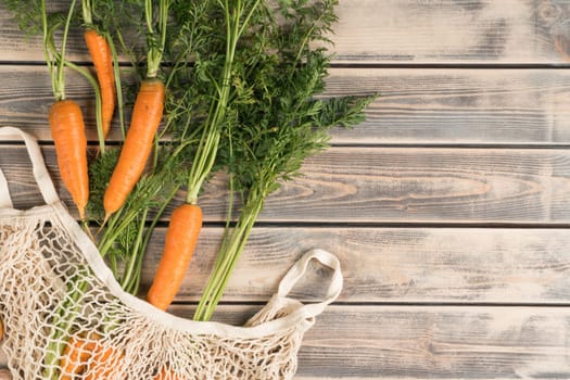 Orange tasty juicy carrot with green stem and leaves in ecological string bag on wooden background. Healthy lifestyle, good vision and carotene vitamins in organic farm food. Top view, copy space