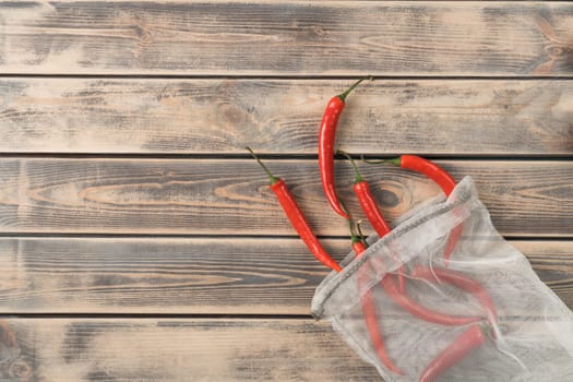 Red hot tasty chili peppers in ecological degradable string bag on wooden background. Spicy food, seasoning and condiment. Recycling and sustainability development, lifestyle. Top view, copy space