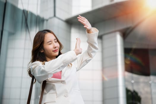 Happy woman wearing white suit jacket with brown bag at sunlight outdoors. Portrait of confidence young businesswoman standing outside office building in city raise your hand to shade sun