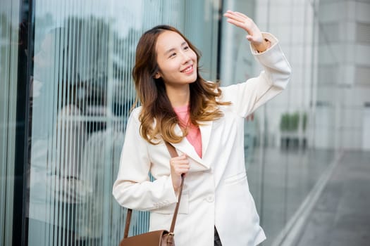 Portrait of confidence young businesswoman standing outside office building in city. Happy woman wearing white suit jacket with brown bag at sunlight outdoors.
