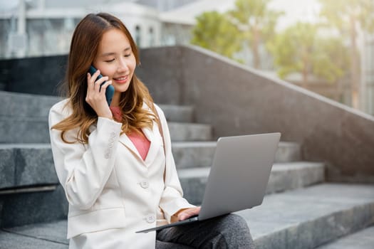 Happy young Asian woman freelance looking laptop computer with smile while talking on mobile smart phone with coffee cup while sitting on stairs outdoors in city. Remote working concept out of office.