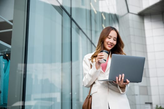Confident businesswoman using computer laptop standing while drinking a cup of coffee at the city. Beautiful smile female being ready to work. Business lifestyle concept.