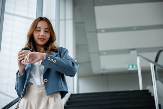Young business woman looking at her wristwatch inside office building and drinking coffee. She wearing blue shirt while go to downstairs in the office.