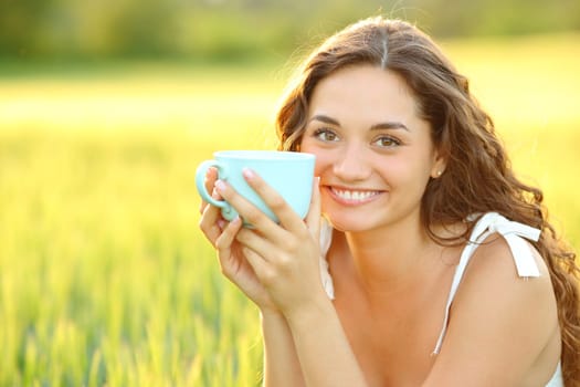 Happy beautiful woman holding coffee cup looking at camera in a wheat field at sunset
