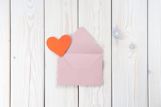Pink opened envelope on white wooden background. Red cardboard heart is taken out of Valentine envelope. Copy space. Love, valentine, confession of feelings. Top view