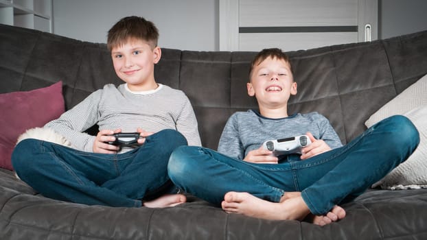 Portrait of two glad satisfied teenage boys sitting on sofa at home, holding gaming controller joystick, playing videogames, having fun. Hobby, free time, gaming, interactive entertainment, leisure.