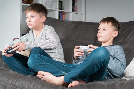 Portrait of two concentrated barefoot teenage boys sitting on grey sofa at home, holding gaming controller joystick, playing videogames. Hobby, free time, gaming, interactive entertainment, leisure.