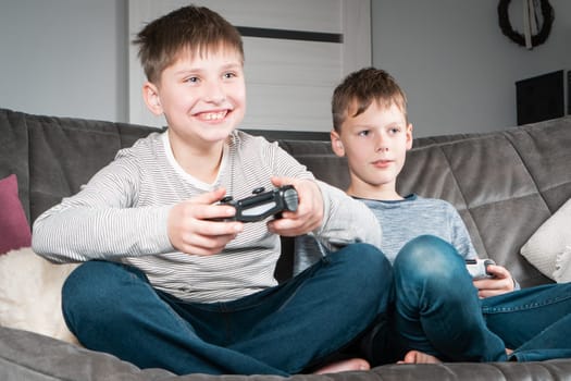 Portrait of two smiling glad teenage boys sitting on grey sofa at home, holding gaming controller joystick, playing videogames having fun. Hobby, free time, gaming, interactive entertainment, leisure.