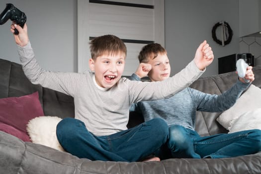 Portrait of two joyful cheerful teenage boys sitting on grey sofa at home, holding gaming controller joystick, playing videogames, celebrating, raising fists, winning competition. Hobby, free time.