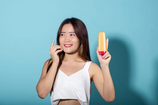 Beautiful Asian young woman holding body lotion on hand in studio shot isolated on blue background, Smiling brunette female hold cream sunblock bottle, tube of sunscreen, protect sun light and UV