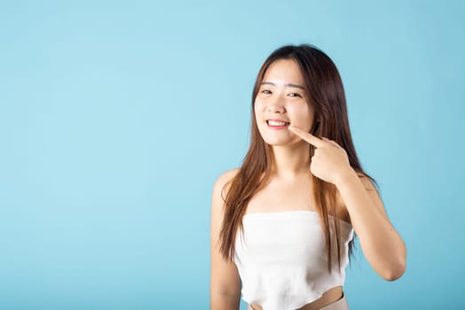 Dental health. Asian young woman pointing to her strong white teeth studio shot isolated on blue background, beautiful female point fingers to smiling mouth