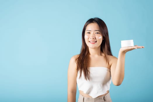 Asian young woman are not very beautiful hold jar of skin cream for face and apply anti-aging moisturizer studio isolated on blue background, Happy female pointing finger her face, pursuit of beauty