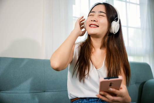Asian young woman wearing headphones listening to music with smart mobile phone and relaxing while sitting on sofa in living room at home, female wear earphone listen music on smartphone