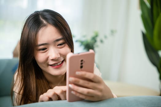 Smiling attractive woman relaxed reading text message on smartphone, Asian young female using smart mobile phone lying on sofa in living room room at home, gadget online application making internet