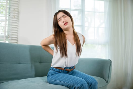 Asian woman holding her lower back feeling painful waist hurt massage in living room, beautiful young female touching back pain sitting in sofa she suffering from backache at home, healthcare medical