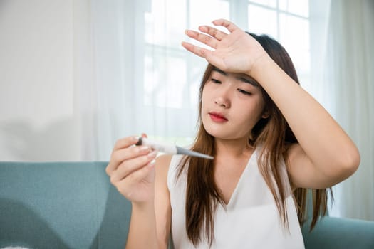 Sick woman have symptom sick high fever she checking her temperature with thermometer in living room at home, young female suffering from flu cold and fever, health care and medical concept