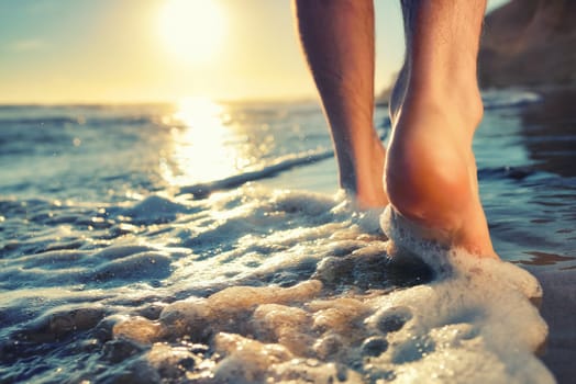 Closeup of a man's bare feet walking at a beach at sunset,with a wave's edge foaming gently beneath them,toned colors