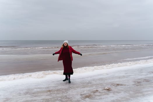 A joyful woman in a red jacket and a dress by the sea in winter.