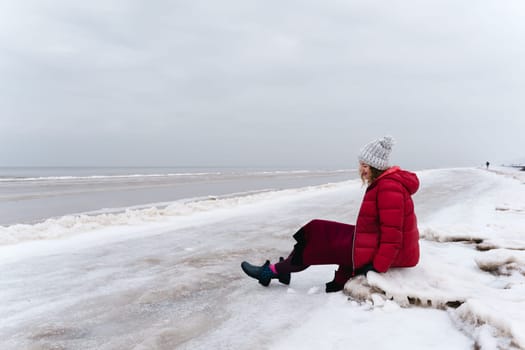 A cheerful woman in a red jacket and a warm dress sits on the shore of the winter sea.