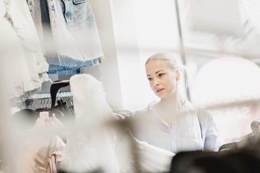 Woman shopping clothes. Female shopper looking at fashionable clothes indoors in clothing store.