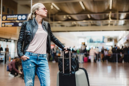 Casually dressed young stylish female traveller checking a departures board at the airport terminal hall in front of check in couters. Flight schedule display blured in the background. Focus on woman.