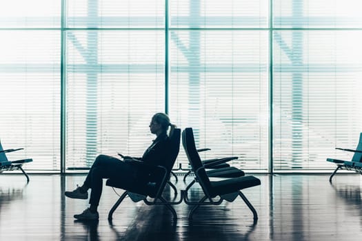 Silhouette of a woman in transit using smarthphone, waiting on airport gate. Horisontal composition. Focus on passanger.