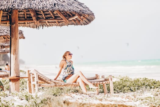 Women sunbathing on dack chair under wooden umbrella on stunning tropical beach. Kiteboarders turquoise blue lagoon of Paje beach, Zanzibar, Tanzania in the background.