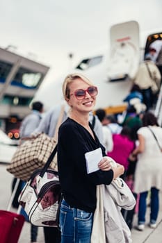 Cheerful woman holding carry on luggage queuing to board the commercial airplane.