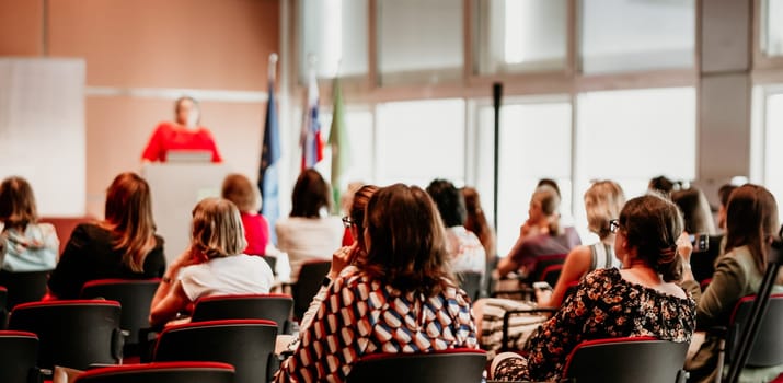 Business and entrepreneurship symposium. Female speaker giving a talk at business meeting. Audience in conference hall. Rear view of unrecognized participant in audience.