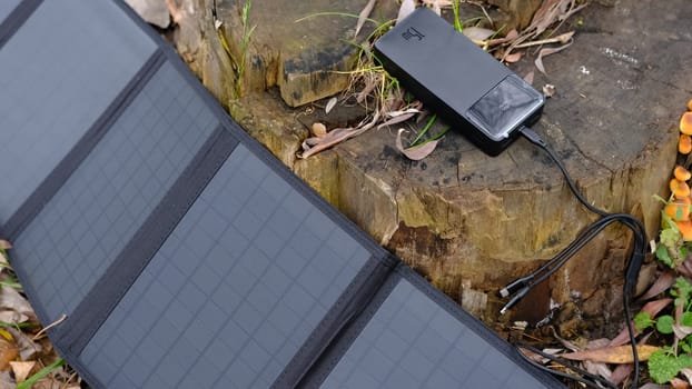 Hands plug in portable solar panel charging power bank by mutlipurpose cable, smartphone, watches, laptops nature background. Clean energy for using in camping or at home when NO electricity