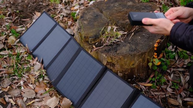 Hands plug in portable solar panel charging power bank by mutlipurpose cable, smartphone, watches, laptops nature background. Clean energy for using in camping or at home when NO electricity