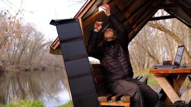Man work on laptop charginf from portable solar panel utdoor in alclove near the river. Clean energy for using in camping. Work with gadgets outdoor with portable solar panels