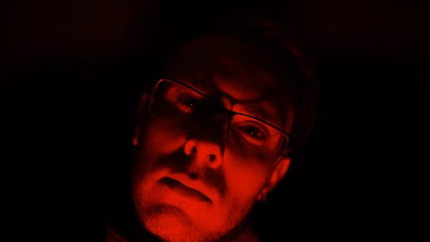the man in glasses looking in camera on a black background in low key anger, psychopath emotions. Horror maniac face in pulsing blue and red light. Mental disorder of mind