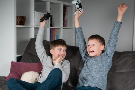 Portrait of two wonderful positive teenage boys sitting on grey sofa at home, holding gaming controller joystick, playing videogames, celebrating, raising hands, winning competition. Hobby, free time.