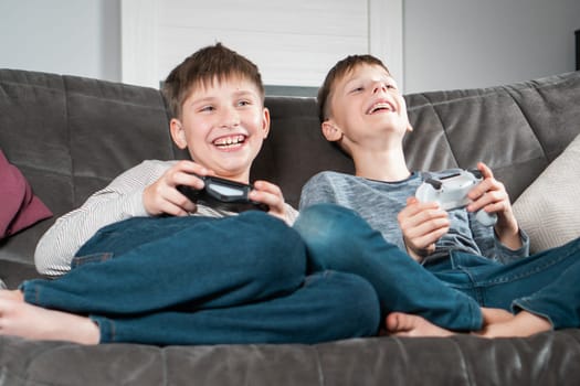 Portrait of two amazing glad barefoot teenage boys children sitting on grey sofa at home, holding gaming controller joystick, playing videogames. Hobby, free time, entertainment, friendship, .