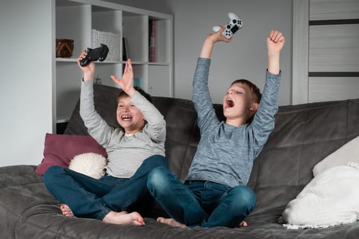 Portrait of two amazing excited teenage boys sitting on grey sofa at home, holding gaming controller joystick, playing videogames, screaming, raising hands, celebrating victory. Hobby, free time.