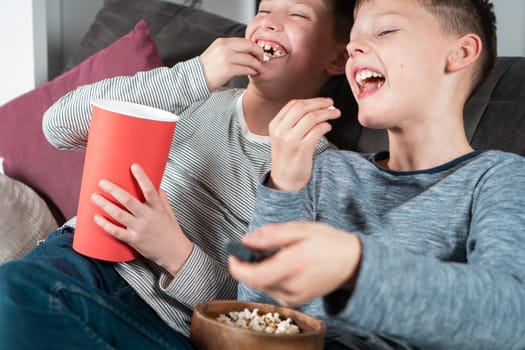 Portrait of two cheerful positive teenage boys sitting on sofa at home, watching funny movie comedy video cartoon on TV, eating popcorn from red bucket, wooden bowl, having fun. Free time, friendship.