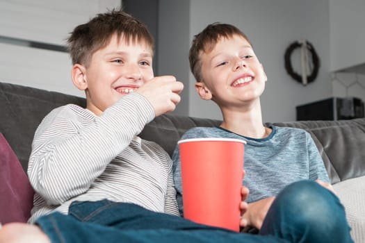 Portrait of two laughing positive teenage boys sitting on sofa at home, watching funny movie comedy video cartoon on TV, eating popcorn from red bucket having fun. Free time, friendship, relationship.