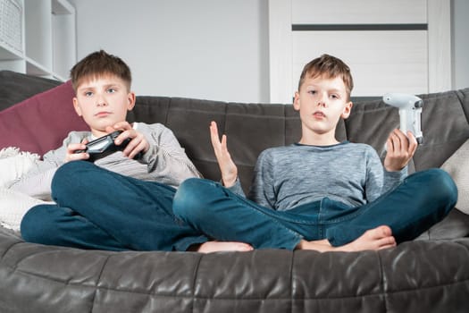 Portrait of two upset displeased teenage boys children sitting on sofa at home, holding gaming controller joystick, playing videogames, raising hand, gesturing. Hobby, free time, gaming, friendship.
