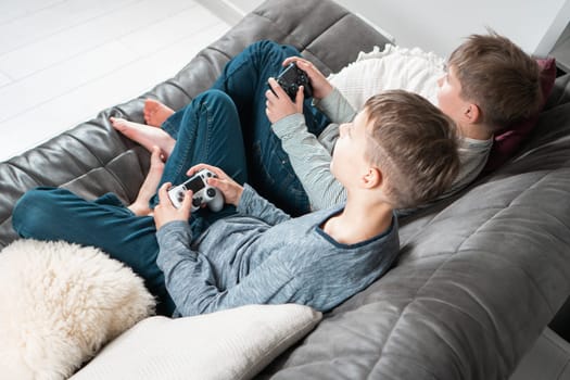 Top view of two teenage boys children sitting on grey sofa near white cushions at home, holding gaming controller joystick, playing videogames. Hobby, free time, gaming, entertainment, friendship.