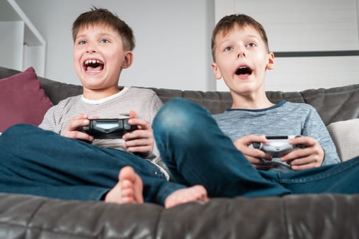 Portrait of two anxious teenage boys children sitting on grey sofa at home with open mouth, holding gaming controller joystick, playing videogames, shouting. Hobby, free time, gaming, entertainment.