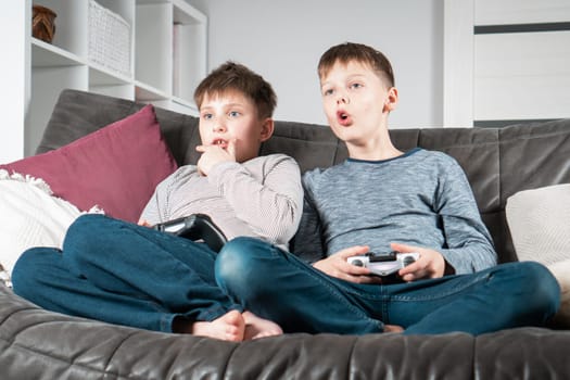 Portrait of two shocked teenage boys children sitting on grey sofa at home with open mouth, holding gaming controller joystick, playing videogames, grimacing. Hobby, free time, gaming, entertainment.