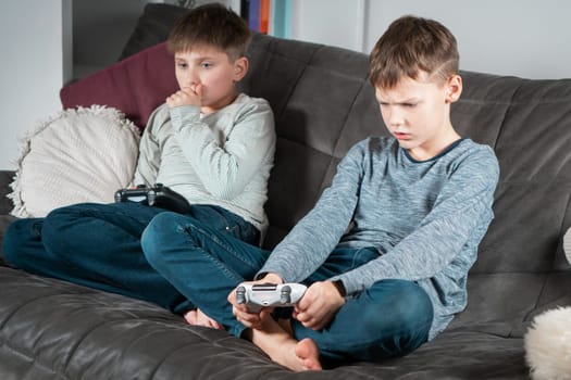 Portrait of two angry indignant teenage boys children sitting on sofa at home, holding gaming controller joystick, playing videogames, covering mouth losing game. Hobby, free time, gaming, friendship.