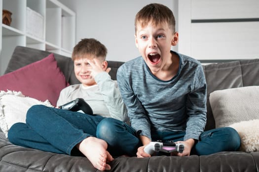 Portrait of two excited teenage boys children sitting on grey sofa at home with open mouth, holding gaming controller joystick, playing videogames, shouting. Hobby, free time, gaming, overemotive.