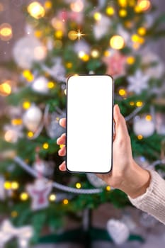 Close up smartphone with empty screen mockup on holiday background with Christmas tree, customer holding phone in hand, shopping online, purchasing gifts, searching information, browsing apps