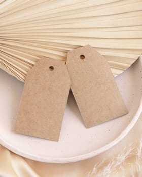 Blank gift tags on beige satin fabric and dried palm leaf close up, mockup. Boho scene with kraft paper envelope. Bohemian or close to nature greeting or wedding Invitation. Romantic composition.