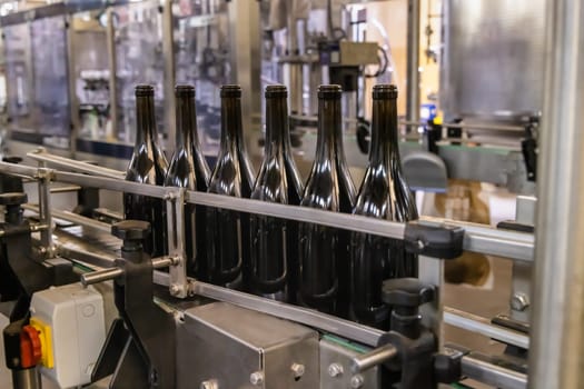 Dark glass bottles being filled with wine by industrial bottling machine at modern wine factory in Italy. Automatic conveyor line for wine-producing