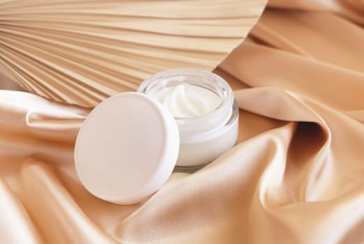 Opened cosmetic jar with white lid on beige satin fabric and dried palm leaf close up, mockup. Boho scene with glass cream jar. Bohemian or close to nature scene.