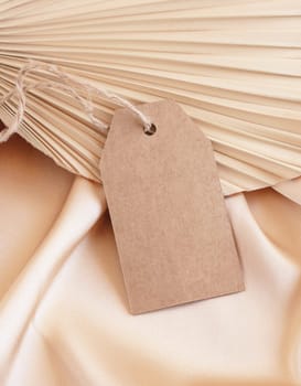 Blank gift tag on beige satin fabric and dried palm leaf close up, mockup. Boho scene with kraft paper envelope. Bohemian or close to nature greeting or wedding Invitation. Romantic composition.